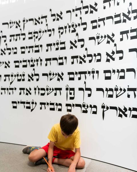 Kind sitzt vor einer Wand mit hebräischer Schrift und schreibt auf einem Zettel.