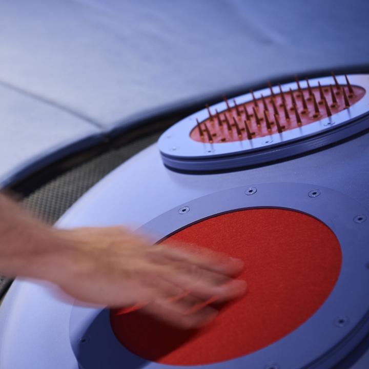 Hand berührt einen roten Kreis, der auf einem blauen Objekt angebracht ist. Im Hintergrund ein weiterer roter Kreis mit stachelförmigen Noppen