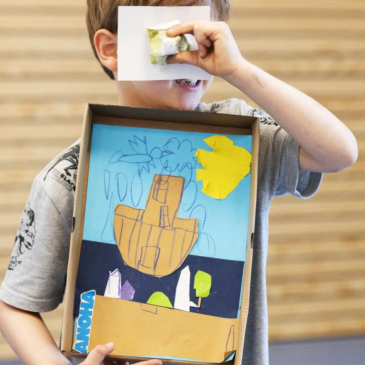 Kind schaut durch ein gebasteltes Fernglas und hält eine Collage mit Schiff im Arm.