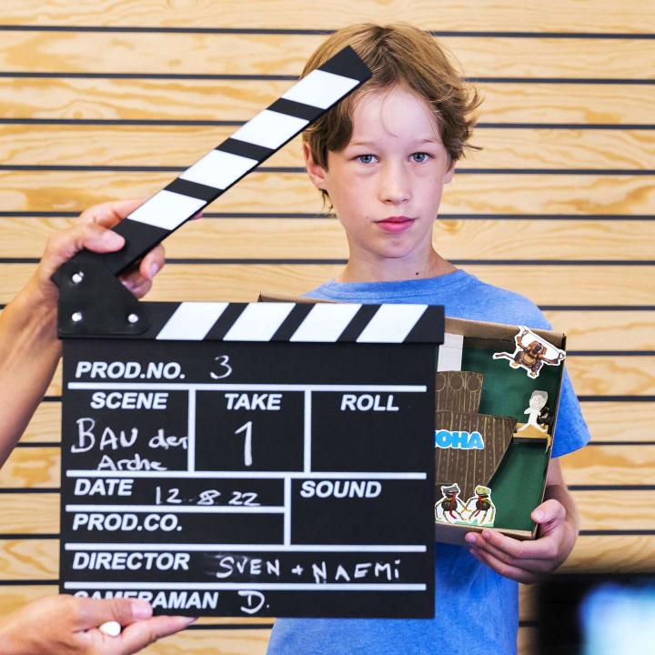 Kind mit eigenem Kunstwerk im Arm schaut in die Kamera. Vor ihm eine geöffnet Filmklappe.
