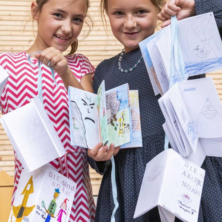 Two children hold self-designed booklets on ribbons and look friendly into the camera.